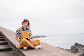 Young woman in a yellow dress reading a book quietly by the sea and taking a break from reality. Concept: disconnection, relaxation, reading