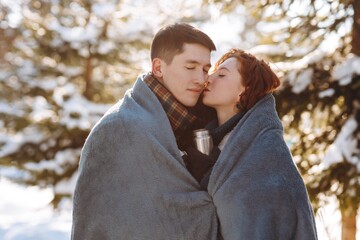 Fototapeta premium Close-up shot of a young couple in love in the middle of a snowy forest. A married couple is standing against the backdrop of snow-covered trees wrapped in a blanket. Young people have a date in