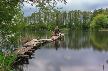 In the early spring morning, a fisherman sits on a long bridge made of planks on the river in complete silence