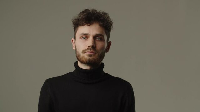 Studio footage of a man with a serious look, looking directly at the camera. A brunette man with curly hair and beard poses against a gray background
