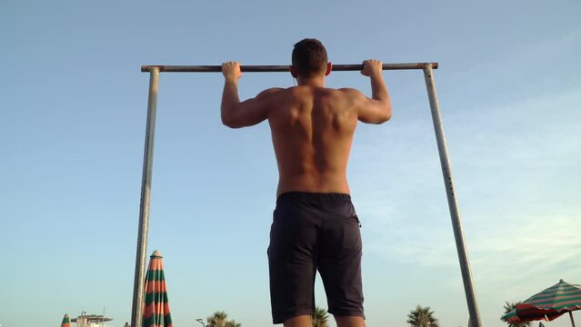 Fit young strong shirtless male athlete doing pull-up on horizontal bar against the blue sky on the hot summer day