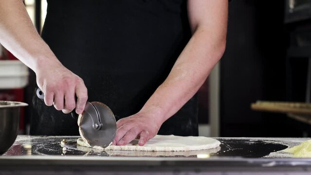 A chef in a professional kitchen trims the edges of a round dough. Preparations for pizza.