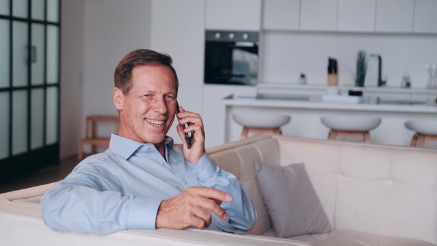 Successful and smiling middle-aged man in blue shirt talking by the phone at home. Sitting on the couch. Living room interior is blurred in background.