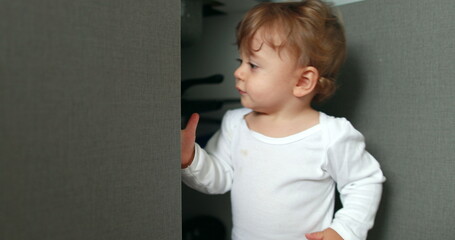 One year old baby boy opening kitchen cabinet cupboard closer door. Infant toddler standing at kitchen floor