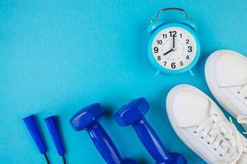 The concept of fitness, sports, weight loss. Top view photo of white sports shoes, blue alarm clock, jump rope and blue dumbbells on isolated pastel blue background