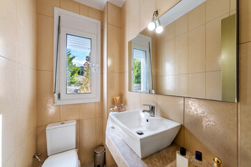 Modern bathroom in light beige colors with white bowl sink, toilet, corner glass shower cabin, tap window, square mirror with light above.