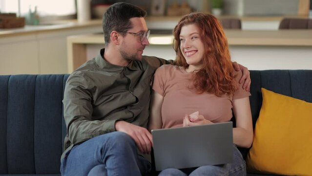 Happy family couple using laptop computer together at home. Smiling husband and wife browsing, surfing, doing online shopping hugging sitting on couch.