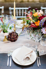 Wedding banquet table setting in blue and white. In the foreground are white plates with napkins, wine glasses, knives and forks. Behind bright flowers. Drapery with tulle.