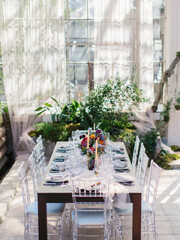 Serving a wedding banquet in the greenhouse. In the center of the table are bouquets of flowers, candles in golden candlesticks, dishes. transparent chairs. Panoramic windows, green plants.