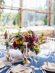 Serving a wedding banquet table in the greenhouse. On a blue tablecloth, a bouquet of bright wildflowers, wine glasses, white plates, cutlery. Behind panoramic windows.