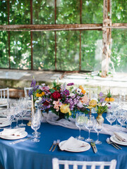 Serving a wedding banquet table in the greenhouse. On a blue tablecloth, a bouquet of bright wildflowers, wine glasses, white plates, cutlery. Behind panoramic windows.