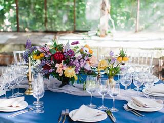 Serving a wedding banquet table in the greenhouse. On a blue tablecloth, a bouquet of bright wildflowers, wine glasses, white plates, cutlery. Behind panoramic windows.