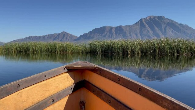 Front end of a small wooden rowing boat floating on a calm water lake with mountains and reeds in the near distance.