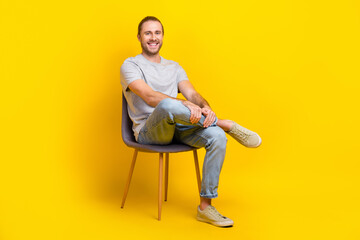 Full size photo pleasant friendly man dressed gray t-shirt denim pants clothes sitting on chair isolated on yellow color background