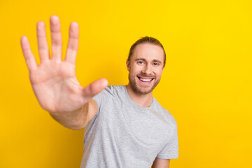 Photo of toothy beaming pleasant man with long hairstyle wear gray t-shirt showing palm at camera isolated on yellow color background