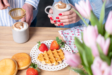 A mans and a woman's hands in light blue clothes with red nails. A man ads cinnamon in a gray cup with caffe latte. The crispy waffle, red strawberries on a white plate and donuts.