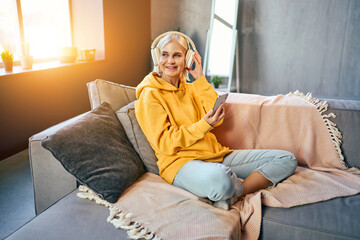 A beautiful senior woman is sitting on a sofa in a yellow hoodie, blue jeans and listening to music in wireless headphones with a phone in her hands and looking away.