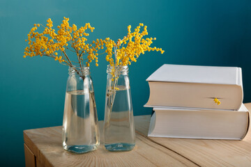 Two beautiful delicate branches of yellow mimosa in small figured glass bottles of water, against a blurry background of a pile of thick books. The concept of spring, reading, details in the interior