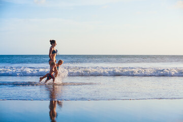 Happy family - mother, kids hold hands and run with fun along edge of sunset sea on sand beach. Active parents and people outdoor activity on tropical summer vacations with children