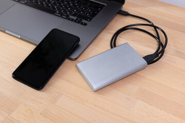 External hard drive with laptop computer and black phone. Portable hard drive connected to a laptop computer on a wooden table. Data transfer or backup between a laptop and a hard disk.