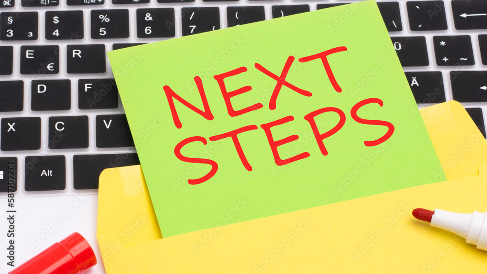 Poster text next steps is written in red marker on a green card. in the frame is a laptop keyboard on yello