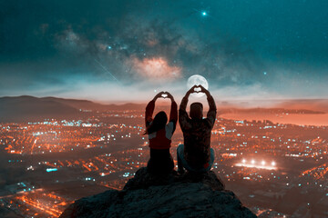 silhouette of a heterosexual couple sitting on top of a mountain making a silhouetted heart shape...