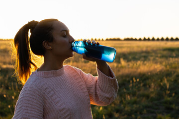 brown-skinned latina woman drinking water from a reusable plastic bottle outdoors at sunset