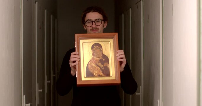 A bespectacled man with a moustache holds an icon in his hands in the corridor. Willingness to bring an object into the light. Focus attention concentration.