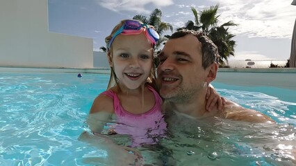 Father and his daughter spend quality time together by the swimming pool at a private villa. They exchange kisses, capturing these precious moments on a wide-angle video selfie.