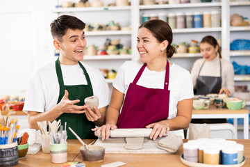 Cheerful young man and woman in aprons modeling handmade mugs of row clay material in pottery workroom