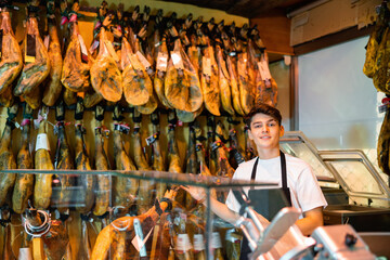 Smiling young salesman in black apron standing near rack with delicious dry-cured jamon legs behind glass counter in butcher shop, offering delicious traditional Spanish meat delicacies.