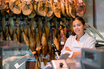 Smiling successful young female butcher shopkeeper standing indoors with crossed arms on background of hanging dry-cured legs of Iberian ham