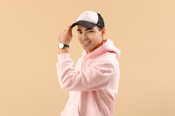 Stylish young man in hoodie and cap on beige background