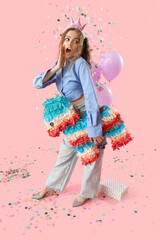 Surprised young woman with pinata celebrating Birthday on pink background