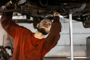 A hardworking mechanic with oil-stained clothing is working underneath the bottom of a car, disassembling and taking off a steering rack.