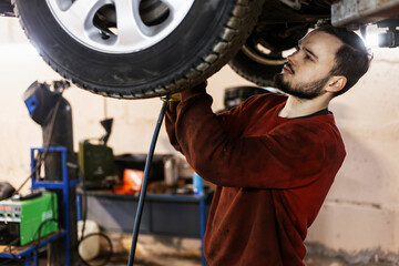 A hardworking mechanic with oil-stained clothing is working with a pneumatic impact wrench underneath the bottom of a car that has been lifted on an electric lift.