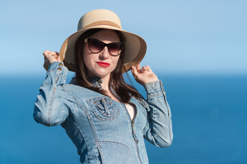 Caucasian ethnicity woman in sunglasses raised her hands and holding straw hat on her head. 40 year old hipster woman in denim jacket standing against blue sky in sunny weather. Summer holiday concept