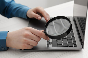 Woman holding magnifier near laptop at wooden table, closeup. Online searching concept
