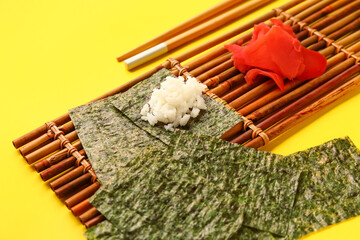 Bamboo mat with nori sheets, rice and ginger on yellow background, closeup