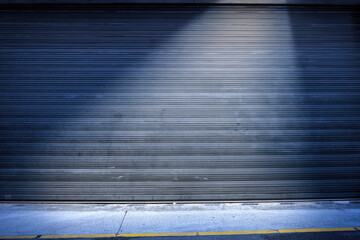 Frontal view of a closed metal roller shutter at night with a sidewalk in the foreground. The empty...