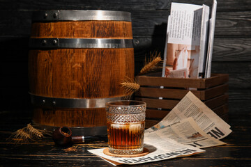 Glass of cold rum with newspaper and oak barrel on dark wooden background
