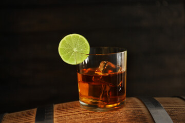 Glass of cold rum on barrel against wooden background