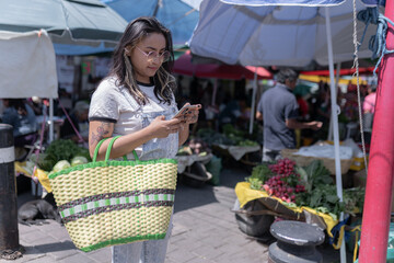 A young Hispanic woman is using her phone in a street market before doing grocery shopping