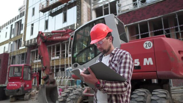 Caucasian man engineer architect holding folder with papers on background excavator, construction machinery at construction site. Builder looking at business plan, architectural documents.