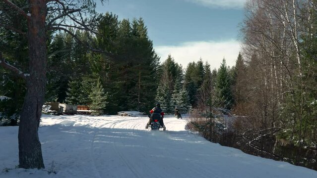 Two snow mobiles driving on a path through the forest on a cold sunny winter day. Static shot.