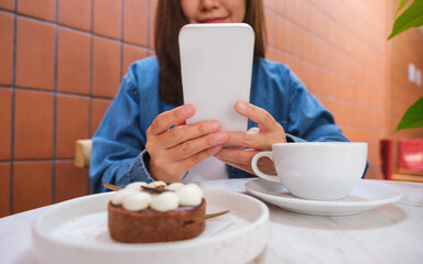 Closeup image of a young woman holding and using mobile phone with cake and coffee cup on the table in cafe