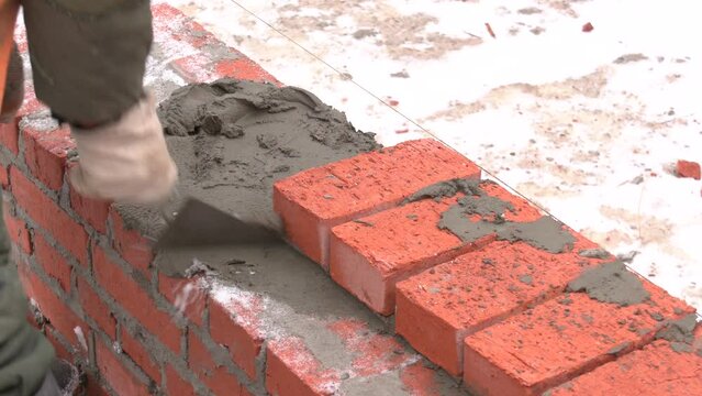 Industrial bricklayer laying bricks on cement mix on construction site close-up. Building a wall of red bricks. Masonry work in details.
