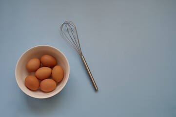 Eggs in white bowl and whisk on blue background