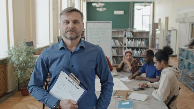 Medium portrait of mature Caucasian man teaching English language in educational center for migrants holding clipboard with grammar tests looking at camera