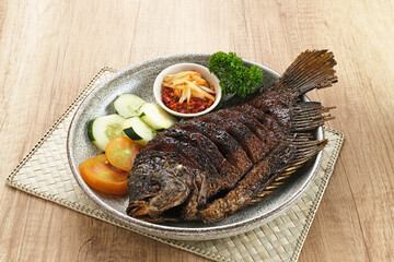 Gurame Bakar Madu, Grilled Gourami with honey and soy sauce. Served with sambal (chilli sauce), cucumber and tomatoes in white plate.   
